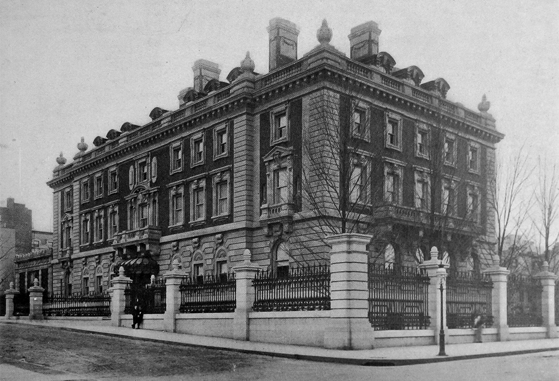 A black and white photograph of a large, rectangular, four-story brick mansion set slightly back from the street corner it's built on. It has rows of windows all along each floor, and a tall, cast iron fence protects it from the surrounding sidewalk. A person stands on the sidewalk, looking miniscule compared to the house towering behind them.