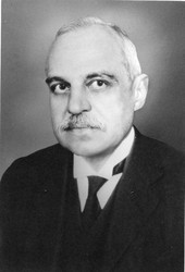 A black-and-white vertical portrait of an older man. He is pictured from the shoulders up and looks directly at the camera. He is wearing a dark three-piece suit and has receding white hair and a moustache.