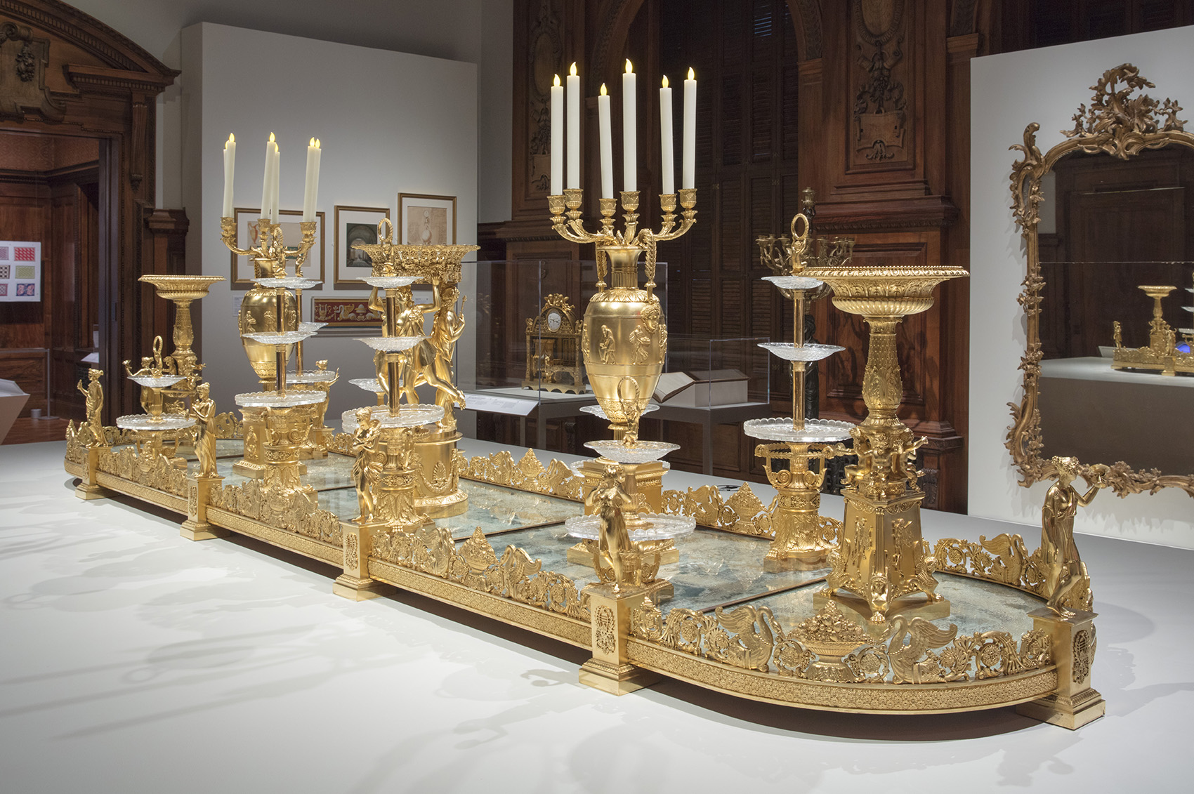 In the center of a gallery, a bronze centerpiece made of 49 individual elements sits upon a platform. The base of the centerpiece is a mirrored plateau made of five sections. On the plateau an array of tiered food display stands fill the space with glass dishes of varied heights. Two candelabra are the tallest elements, lit dimly with a flickering light.