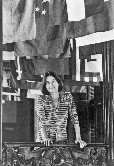 Black-and-white photograph of a woman looking over an ornately carved wooden banister. A series of flags hang from the ceiling behind her.