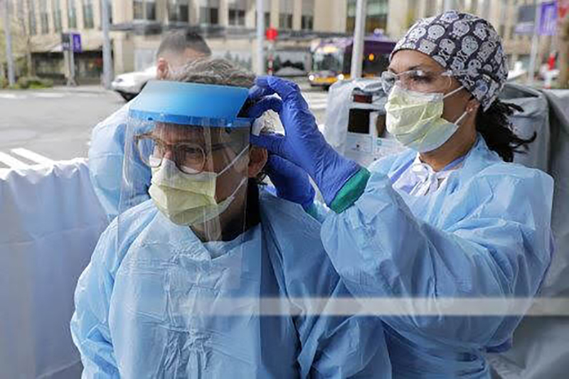 A doctor in scrubs wearing a paper face guard adjusts a face shield worn by her colleague, who is also in scrubs and wearing a paper face shield. The face guard is a headband to which is attached a sheet of clear plastic that covers the entire face