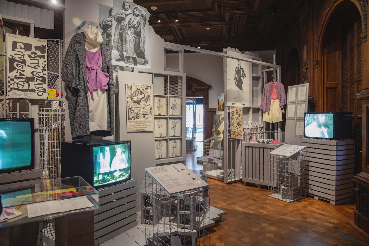 Outfits, videos, and ephemera hung on gray structures within a gallery space