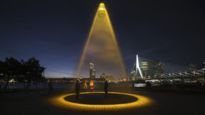 A night-time photograph of a public space with a high suspended light source which illuminates a large yellowish open circle on the paved surface below.
