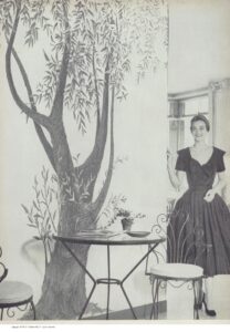 A woman in a dress stands beside a wall decorated with a large mural of a tree and before it are two chairs and a table.