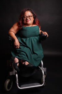 Photo of Olivia M. Asuncion, a Filipina woman with dyed-red hair wearing a rich green dress and elegant blakck heeled shoes. She wears black framed glasses and sits in a power wheelchair.