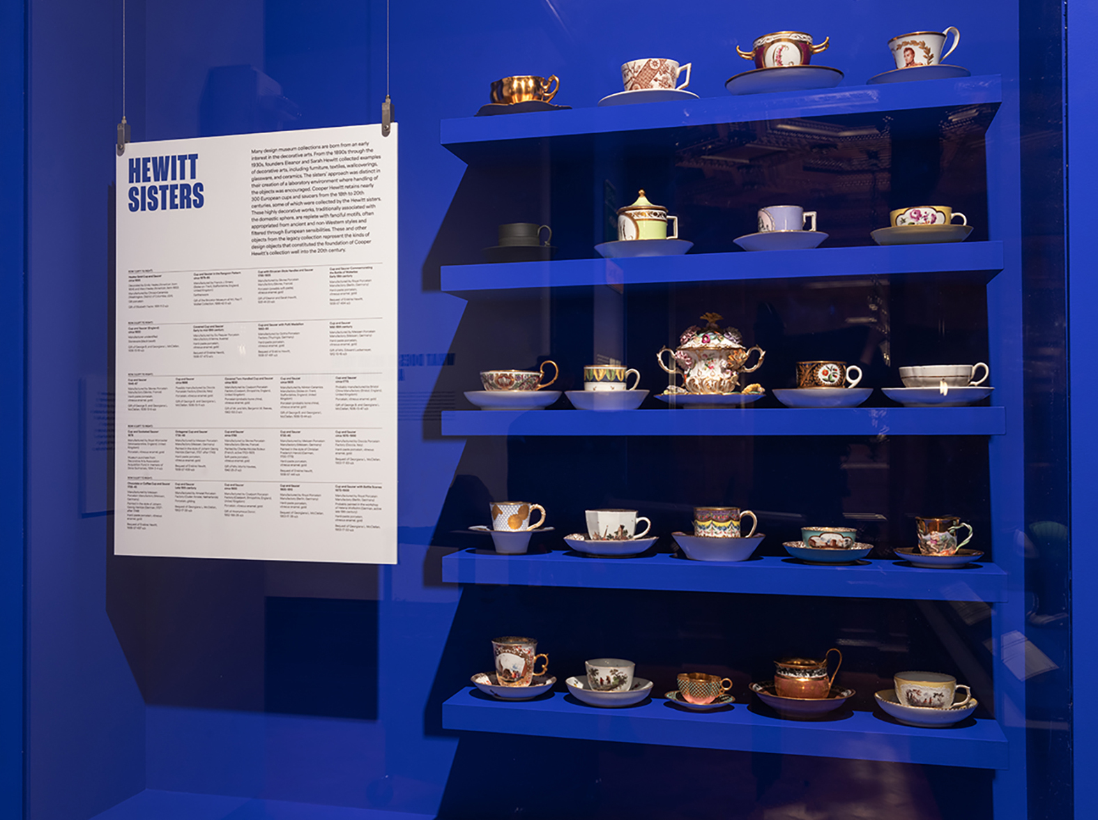 Against a vibrant blue wall, five shelves are installed, each holding either four or five ornately decorated and gilded cups and saucers.