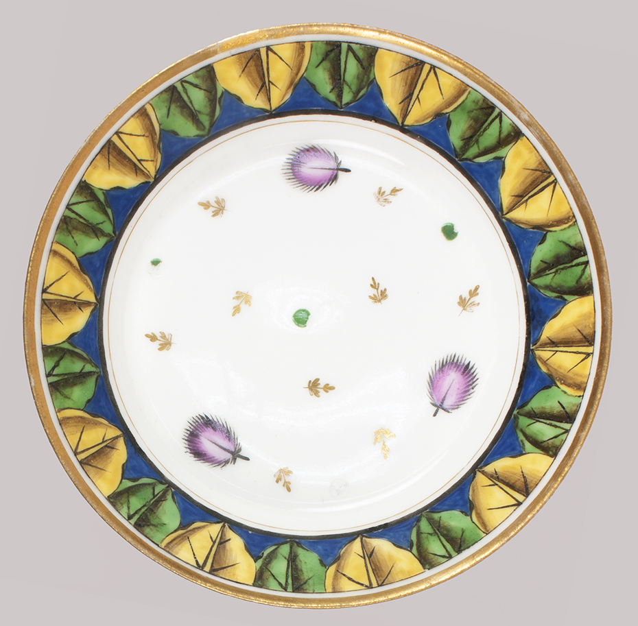 Overhead view of a saucer with a gold rim, band of alternating yellow and green leaves against a blue band, and, at the center, a sparse pattern of pink leaves and gold feathers.