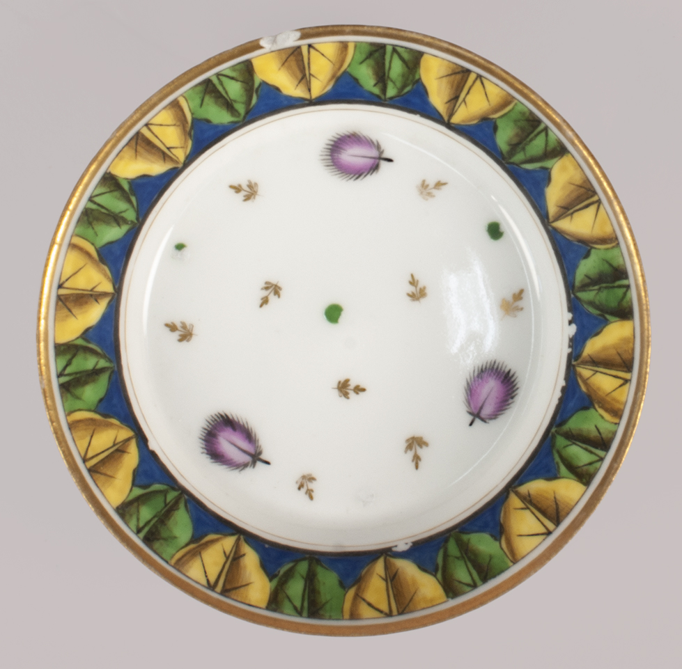 Overhead view of a saucer with a gold rim, band of alternating yellow and green leaves against a blue band, and, at the center, a sparse pattern of pink leaves and gold feathers.