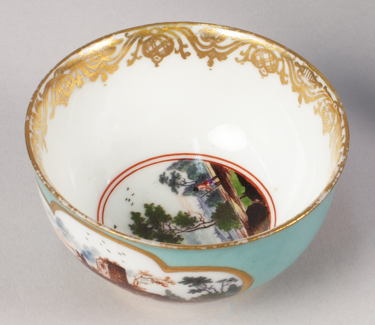 Overhead view of a teacup that is teal on the outside and white on the inside. The rim boasts a decorative gilded pattern and pastoral scenes of featured in vignettes on both the cup’s side and interior bottom.
