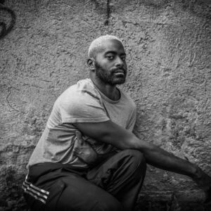Black and white photo of Jerron, a dark skinned black man with a beard and cropped blonde hair, seated in front of a slightly tagged concrete wall, his arms lightly crisscrossing over themselves as he stares directly at us.
