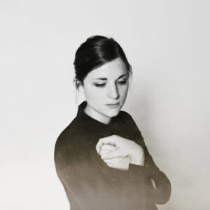 Black and white photo of Molly, a white female with brown hair in a bun. She is wearing a black dress and her hands are clasped together against her chest.