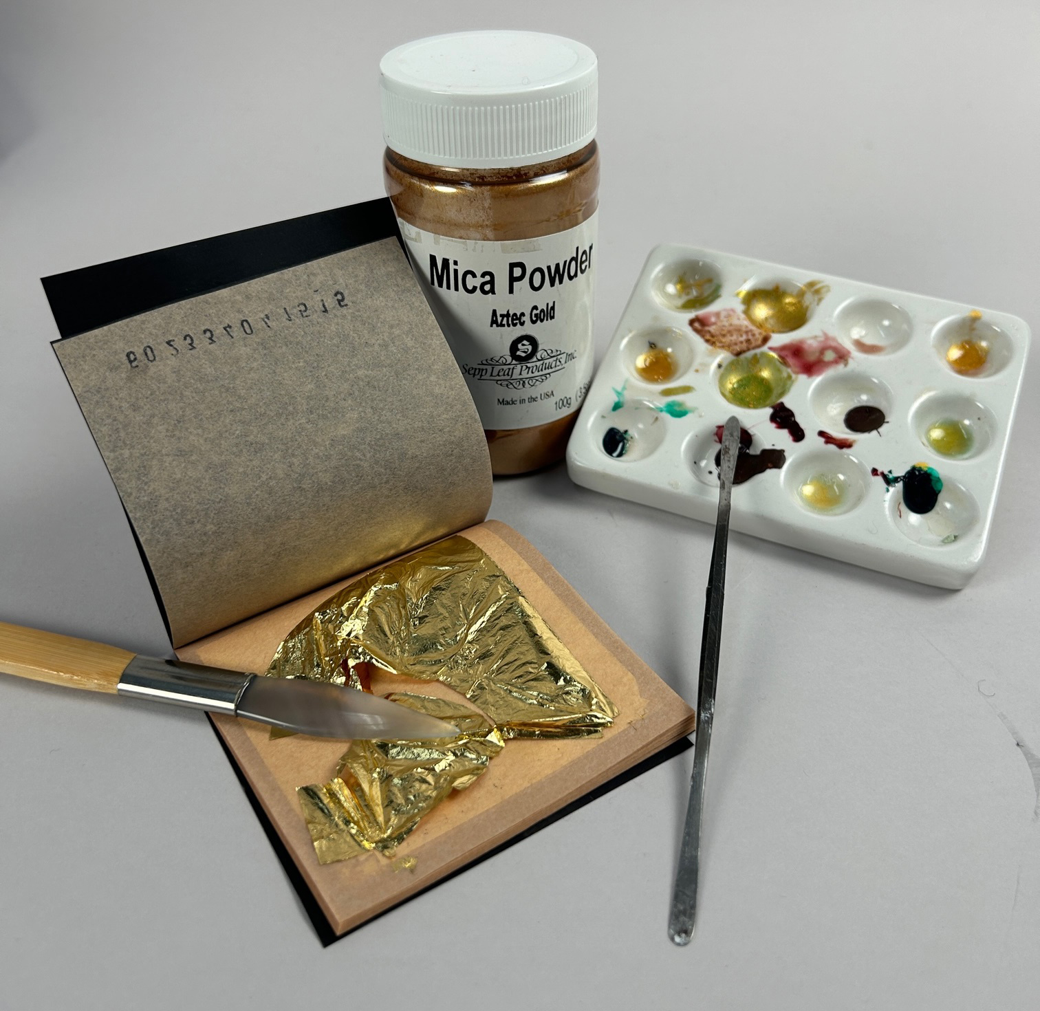 On a blank surface is a pad on white rests gold leaf, a jar containing a gold substance and displaying the words “Mica Powder / Aztec Gold”, a paint palette with samples of a variety of gold and black hues, and two spindly tools.