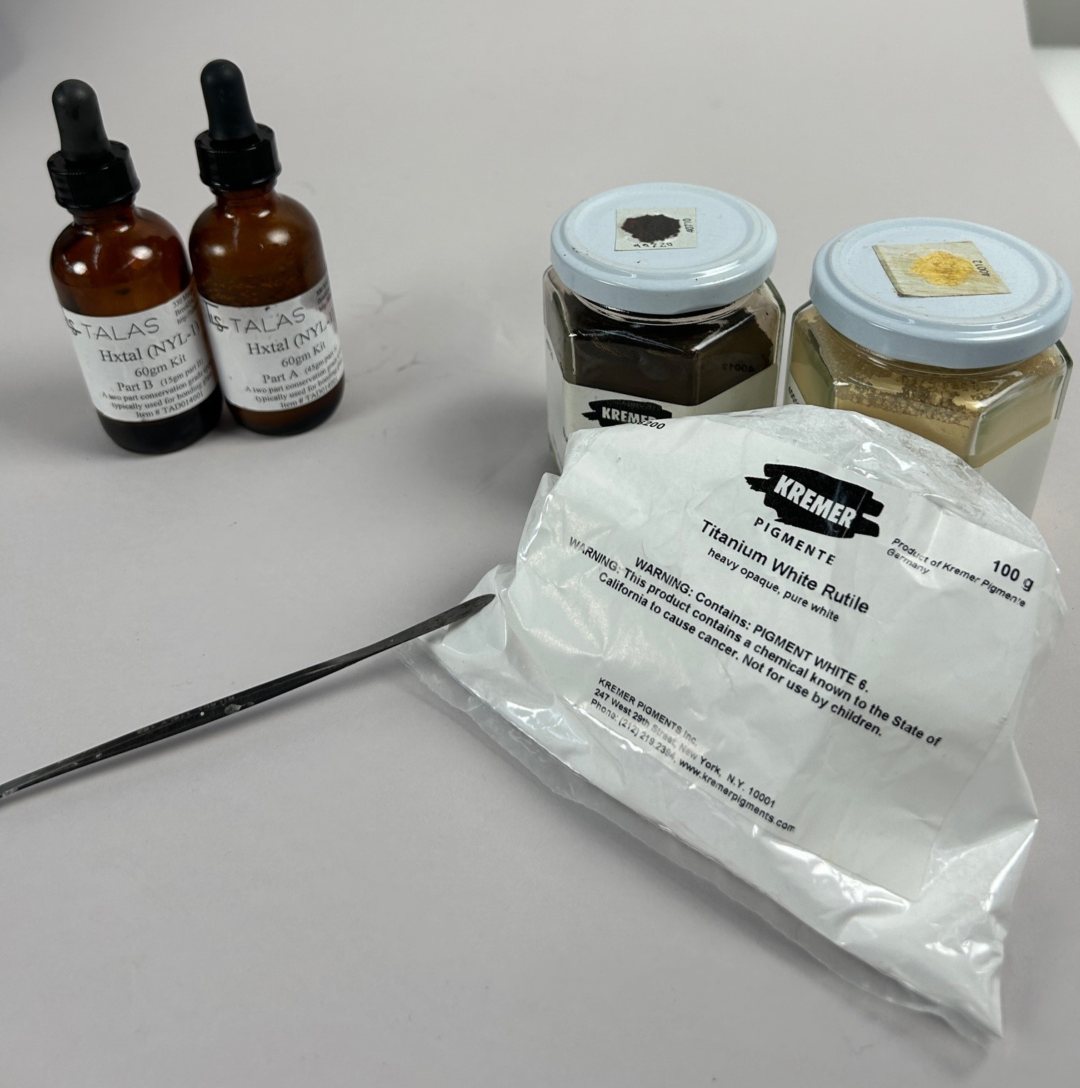 On a blank surface are two small bottles of chemicals, two jars of pigment (both black and gold), a scalpel, and a plastic bag containing a white substance.