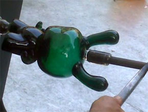 Emerald glass blown object in the shape of a robot. A light-skinned hand holds the end of a punty rod that is attached to the head of the object.