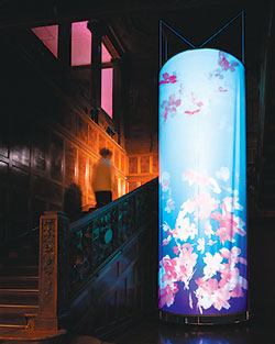 Photograph of a 2-story glowing, lantern-like blue cylinder with pink cherry blossoms inside next to the dimly lit paneled wood staircase of Cooper Hewitt’s front hall. A blurry image of a person walks up staircase at mid-height of the cylinder, and diffused rose-colored light glows through a window on the visible second story.