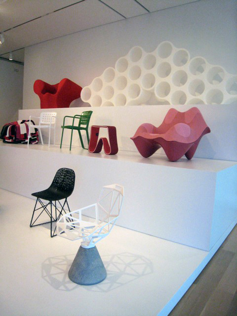 Angled photograph of showroom displaying a three-tiered assemblage of modern, contemporary, and non-traditional styles of seating. Each white tier displays two to five unique, experimental designs of seating in solid red, white, green, pink, grey and black colors. Chairs are in various playful shapes and forms.