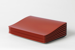 Bright red minimal rectangular form; slightly undulating top panel and accordion-like vents visible from three sides; low, black rectangular base.
