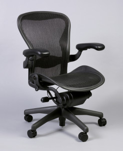 Task chair of black mesh stretch fabric within black frame consisting of contoured back, broad contoured seat with sloping front edge, adjustment mechanism under seat; black, adjustable padded arm supports; base consisting of vertical post on five radiating feet with casters.