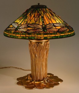 Conical shade of amber and green stained glass with dragonflies around the sides, on gilded base as bunch of tall grass surrounded by lily pads.