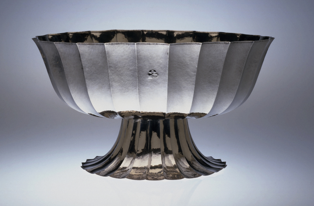 A fluted oval raised silver fruit bowl with matte finish, engraved with a monogram on the sides, all on a with a raised fluted oval foot that splays out from the bowl.