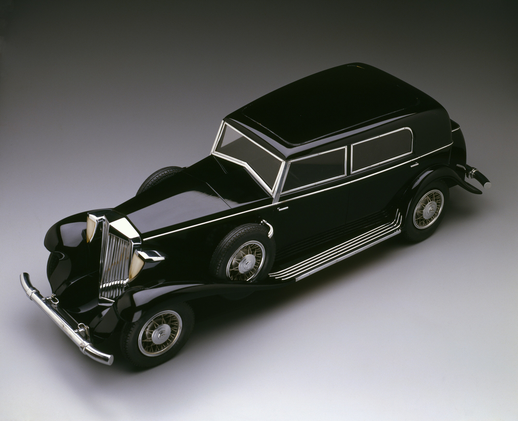 Black sedan with chromed grille, headlights, bumpers, running boards, window frames, and door handles; rubber tires with chromed hub caps; two spare tires mounted on either side, at front fenders. Model mounted on flat wooden base with metal plaque mounted at front. Removable plexiglass and wood cover fits on base.