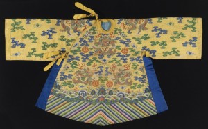 Small robe in yellow silk tapestry weave, k'ossu, with metallic gold dragons on the front, back, shoulders and sleeves. The ground is filled with clouds motifs in blue and green; flaming pearls, bats, and other auspicious symbols. At the bottom is a deep border is multicolored diagonal stripes (water convention). Assymetrical closure with yellow silk ties at the neck and side; side openings are edged with bright blue silk.