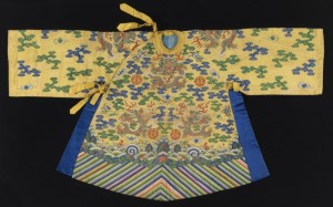 Small robe in yellow silk tapestry weave, k'ossu, with metallic gold dragons on the front, back, shoulders and sleeves. The ground is filled with clouds motifs in blue and green; flaming pearls, bats, and other auspicious symbols. At the bottom is a deep border is multicolored diagonal stripes (water convention). Assymetrical closure with yellow silk ties at the neck and side; side openings are edged with bright blue silk.