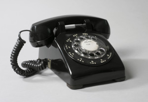Black rectangular plastic body with sloping top; circular clear plastic dial in center, surrounded by white numbers 1 to 0, the letters A to Y, and the word "OPERATOR", all arranged in a circle around the dial, their positions corresponding to fingerholes in dial. Barbell-shaped handset of black plastic sits in cradle on top of body; coiled black wire at one end of handset plugs into telephone body.