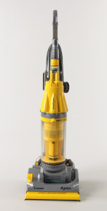 Upright vacuum cleaner: metalic gray, yellow, and clear housing leading up to handle, with cavities for tools (brushes, tubes); front dominated by motor and collection bin assembly of yellow and clear plastic; at bottom, wide, low cleaning head with brush in front, two wheels in back, yellow at joints and adjustment points.