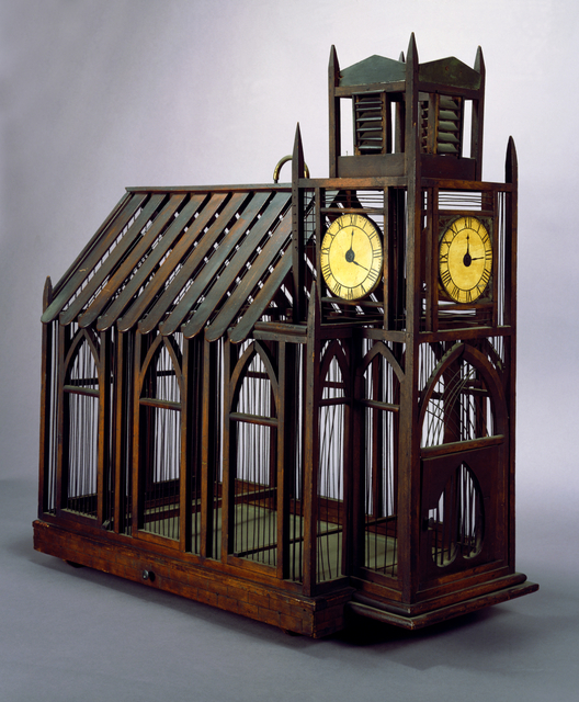 Birdcage of wooden slats, dowels and metal wire in the form of a church with peaked roof; walls of clerestory-like gothic style arches; at one end a rectangular bell tower with three large, circular, painted clock faces, and door with inverted heart-shape perforation. Wood and sheet metal under tray (b) pulls out at left side. Small loop handle of brass on roof.