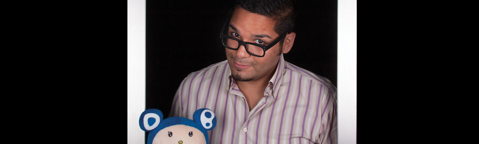 Bespectacled man standing inside a picture frame holding a blue stuffed bear.