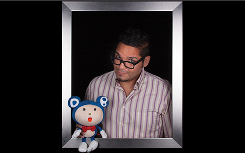 Bespectacled man standing inside a picture frame holding a blue stuffed bear.