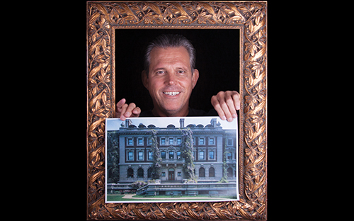 grinning man standing inside an ornate picture frame holding a photo of a mansion.