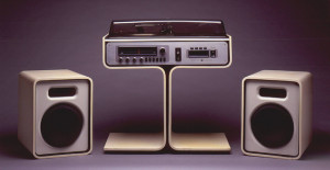 Unit comprising record turntable, radio/tuner and eight-track casette player in rectangular housing in top of white enameled wood pedestal (a); turntable and casette storage well in top, control knobs and radio on front; rectangular tinted plastic lid (b). Two upright rectangular speakers of white enameled wood faced with white plastic and dark fabric (c,d); speakers can fit on rectangular pedestal feet on either side of center column.