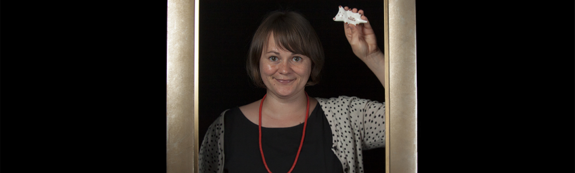 Brown-haired woman standing inside a picture frame holding a small ceramic cat in her right hand.