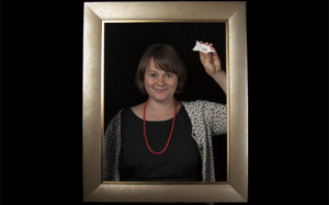 Brown-haired woman standing inside a picture frame holding a small ceramic cat in her right hand.