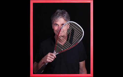 Man standing inside a picture frame holding a squash racket over one eye.