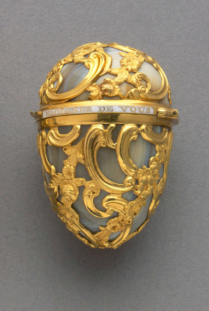 Egg-shaped form of curving rococo gold cage work over gray agate. Hinged lid with white enameled band showing the phrase, "Eloignez de vous rien n'est agreable" (Separated from you nothing is pleasant).