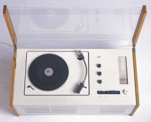 Rectangular form; white metal housing flanked by ash wood sides; turntable and arm on left, control knobs and radio tuner on the right. Hinged cover of clear plastic with ash wood strips on sides.