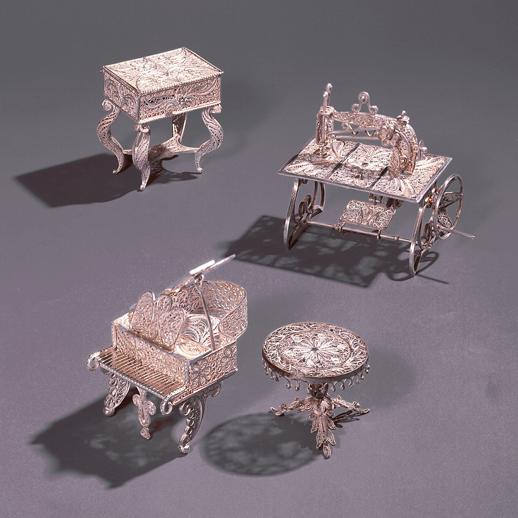 Filigree miniature comprising sewing machine form on rectangular table top with decoration in three panels, on scrolled supports. The working parts consist of a treadle, crank, slotted arm, and needle