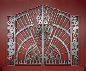 Pair of gates, each a mirror image of the other; gate -1 with handle. Each gate with geometric decoration in two sections: a tall narrow rectangular section at outer left of gate -1, and outer right of -2, hinged to a larger trapezoidal section. The narrow rectangular sections with decoration of 14 irregularly-stacked cogwheels. The trapezoidal sections with radiating arcs, lightning bolt-like forms and zigzag lines emanating from lower inside corners; cogwheels, spirals, and other shapes interspersed along inside edges; bottom edges with segmented border depicting stacks of coins.