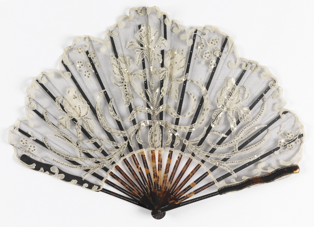 Folding fan with a black silk net leaf with white cotton needle lace motifs of irises, scrolls and flower sprays applied; lace elements embroidered with silver -colored steel spangles. The sticks are of tortoise shell, piqué, with guards ending in a serpentine form; ebony slips; glass stone at the rivet.