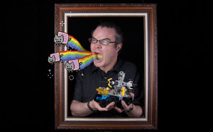 man holding a big lego structure and standing inside a picture frame. Pixel art of rainbow nyan cats are photoshopped to be flying out of his open mouth.
