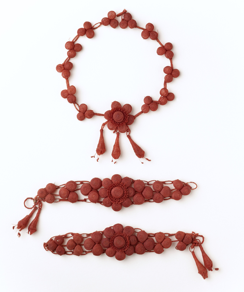 A red colored necklace made of weaved balls in threes, with a centered flower, dangling three tear drop shapes. Bracelets are red colored and made of weaved balls, with a centered flower, dangling two tear drop shapes at the end.