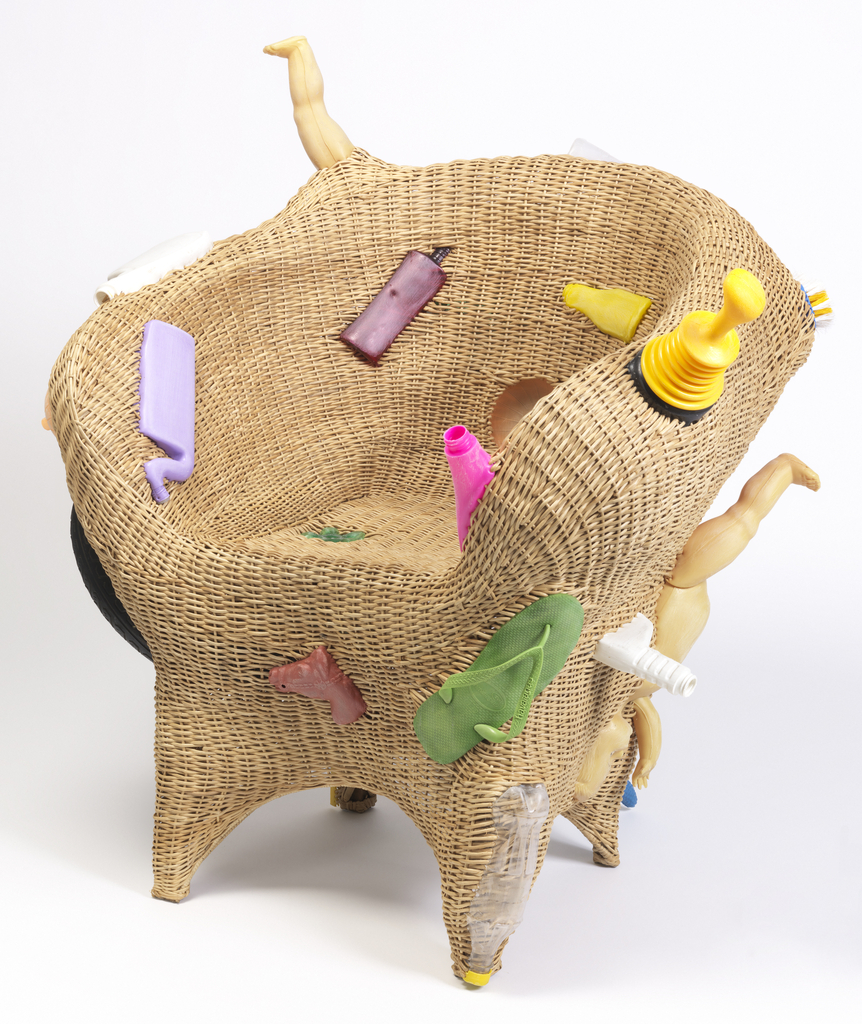 Woven tan wicker armchair with colorful found plastic and rubber objects (including discarded bottles, tire, flip-flop, broken doll parts) woven into, and protruding from, the form.