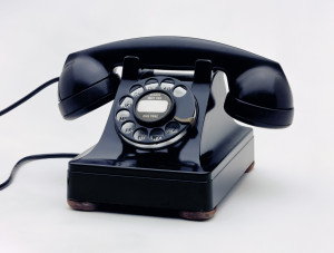 Black rotary telephone; transmitter/receiver handset sitting in cradle on raised body with square base; black metal finger wheel above white dial with black numbers; smooth cord, one end attached to handset transmitter and the other to back of telephone base.