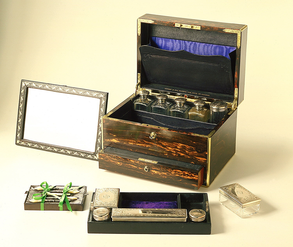 rectangular box,lined with purple silk, fitted with leather document holders, and mirror with gold-leaf foliate border, the upper level withy 4 rectangular and 1 central faceted silver-lidded cylindrical cologne jars, 3 silver-lidded glass boxes, 2 silver-lidded glass ink bottles, mother-of-pearl handled sewing implements, tweezers, button hook, pen knife and hole punch or scorer-all in the upper compartment, a secret drawer below fitted with a letter writing slide.
