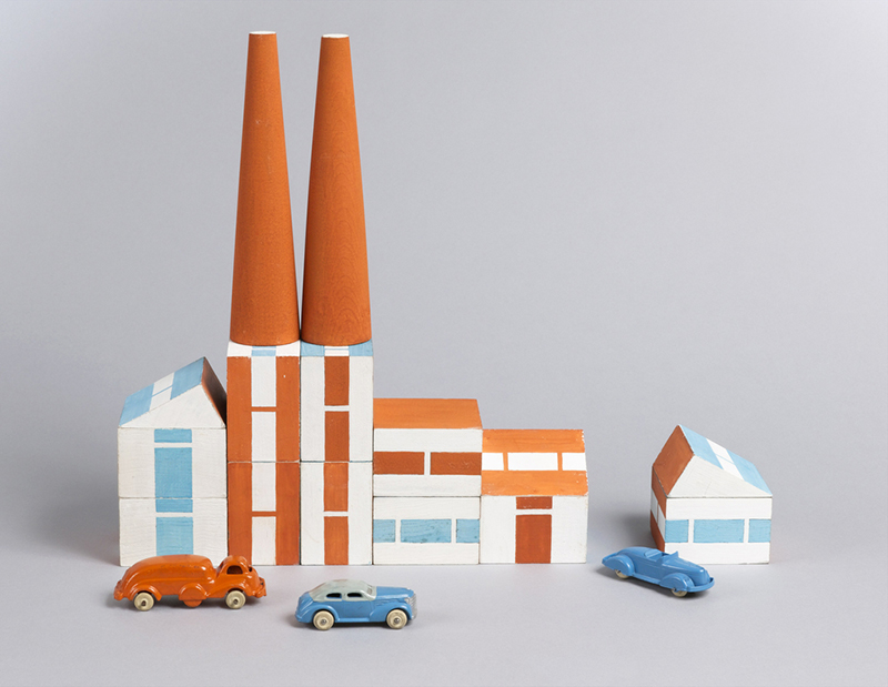orange, white and blue geometric blocks stacked up to look like a row of buildings against a gray backdrop with toy cars in front.