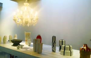 A large wooden table with a sparse arrangement of ceramic cups and vases on top, as if in a fancy shop, and an unusual chandelier dangling over the table, which has the shape of a traditional candle chandelier but the surface of the chandelier is completely coated in tiny white crystals, almost like an even coating of barnacles.