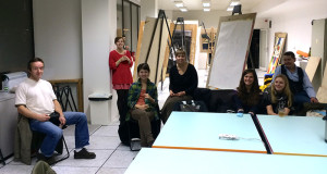 A group of young white people casually gathered around a large empty work table. Easels in the background. It is night.